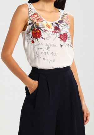 Wit mouwloos topje met kleurrijke bloemenprints en handgeschreven tekst aan de voorkant, gecombineerd met marineblauwe wijde broekspijpen.