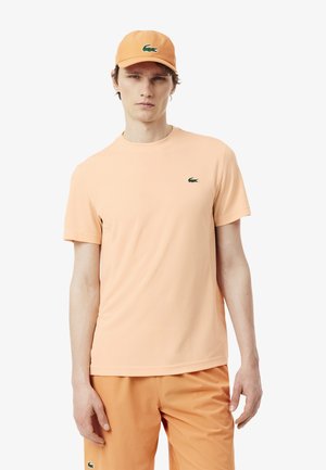 Mann trägt ein pfirsichfarbenes T-Shirt, passende Shorts und eine Kappe, jeweils mit einem kleinen grünen Krokodil-Logo auf der Brust, am Bein und vorne auf der Kappe.