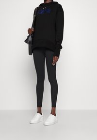 Felpa nera con logo blu, abbinata a leggings neri e sneaker bianchi. Tessuto morbido, vestibilità comoda e cappuccio con coulisse.