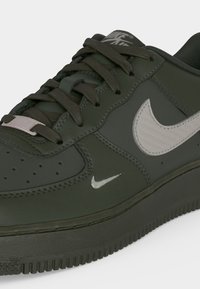 Σκούρο πράσινο αθλητικό παπούτσι Nike Air Force 1 με λευκά σημάδια Nike, μαύρα κορδόνια και υφή στη σόλα, απεικονιζόμενο σε κοντινή λήψη από πλάγια και επάνω γωνία.