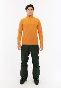 Oranžový fleece pulóver s dlhým rukávom a polovičným zipsom pri golieri. V kombinácii s tmavozelenými nohavicami, čiernymi lyžiarskymi topánkami a minimálnym brandingom.