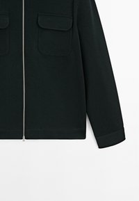 Veste noire zippée en tissu texturé, présentant deux poches avec rabats et un détail de manche à revers au niveau de l'ourlet.