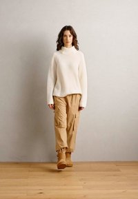 Crème ribbel turtleneck trui gecombineerd met beige cargobroek. Model draagt tan laarzen. Eenvoudig ontwerp met een relaxte pasvorm en minimale accenten.