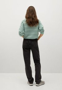 Personne aux longs cheveux bruns, de dos, portant un pull vert clair texturé, un jean noir et des baskets grises sur un sol blanc.