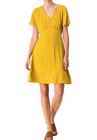 Femme portant une robe jaune à manches courtes avec des pois blancs, col en V, boutons et sandales marron à bout ouvert, debout sur un fond blanc.