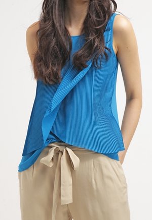 Kobieta ubrana w niebieską plisowaną bluzkę bez rękawów z asymetrycznym przodem oraz beżowe, wysokie spodnie z tkaninowym paskiem wiązanym w pasie.