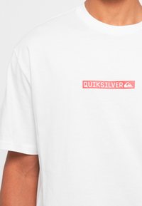 Person wearing a white T-shirt with a small red Quiksilver logo and text on the left chest area.