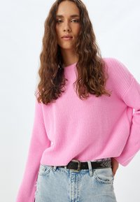 Rosa stickad tröja med en lös passform och ribbad struktur, parat med ljusblå slitna jeans och ett svart skärp med nitar.