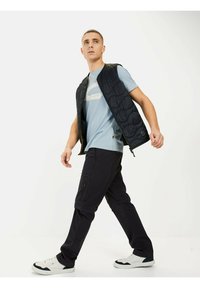 Sort quiltet vest over en lyseblå t-shirt, parret med mørke cargo-bukser og hvide sneakers. Vesten har en lynlås og en moderne pasform.