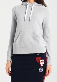 Sudadera gris hecha de un tejido suave, con un bolsillo canguro en el frente, cordones ajustables y puños acanalados. Llevada sobre una falda navy con parches.