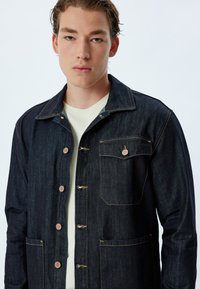 Denim jacket in dark blue with copper buttons, two front pockets, beige stitching, and a classic collar. Worn over a light-colored shirt.