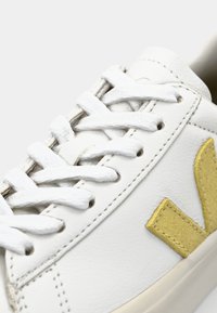 Vita lädersneaker med texturerat tyg, platta vita snören och en gul V-formad detalj på sidan. Sytt detaljer finns.