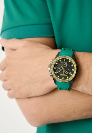 Orologio nero e oro con cinturino in gomma verde. Presenta un quadrante testurizzato, più quadranti e una scala tachimetrica sulla lunetta.