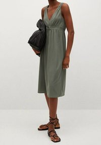 Oliwkowa sukienka bez rękawów z dekoltem w serek, wcięciem w talii i długości do połowy łydki. Noszona z czarną fakturowaną torebką i sandałami na ramiączkach.