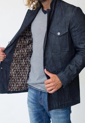 Mann trägt eine blaue Denimjacke mit geometrisch gemusterter brauner Innenseite, kombiniert mit einem hellgrauen Hemd und blauen Jeans.