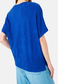 Top corto de manga corta azul en una tela suave con un patrón floral texturizado, escote redondo y un corte relajado. Combinado con unos vaqueros de mezclilla azul claro.