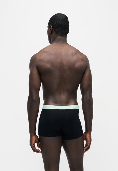 Homme athlétique debout, dos à la caméra, portant un boxer noir avec une ceinture claire sur fond blanc uni.