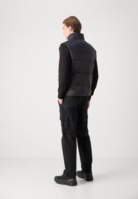 Gilet imbottito nero con colletto alto, abbinato a pantaloni cargo neri e scarpe scure. Il gilet presenta sezioni trapuntate e una texture liscia.
