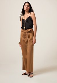 Camisola nera con pizzo abbinata a pantaloni beige a vita alta con cintura da annodare. La modella indossa sandali neri e una collana di perline.