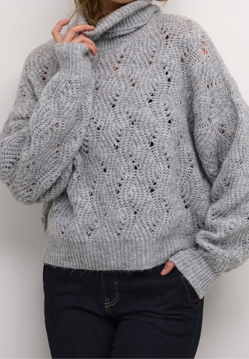 Personne portant un pull gris à col roulé en maille torsadée avec un motif texturé, associé à un jean bleu foncé, la main touchant le cou.