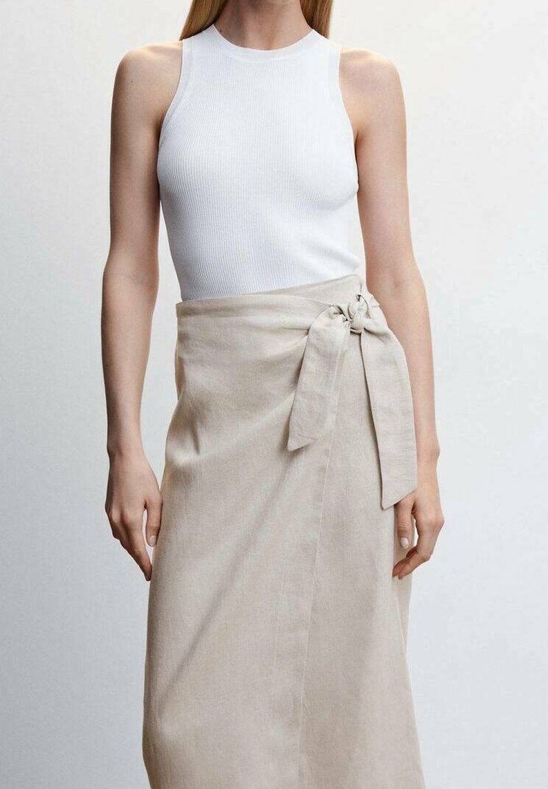 Femme portant un haut blanc côtelé sans manches et une jupe beige portefeuille nouée à la taille, debout devant un fond clair uni.