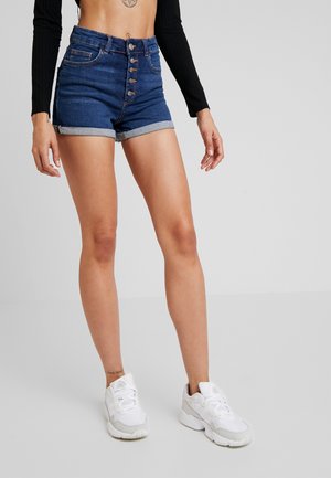 Shorts en jean bleu foncé à taille haute, fermeture à cinq boutons sur le devant, revers roulés et deux poches avant. Portés avec des baskets blanches.