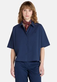 Maglietta a maniche corte blu navy con bottoni, dal taglio leggermente corto, caratterizzata da una sottile trama e un piccolo logo sul lato sinistro del torace.