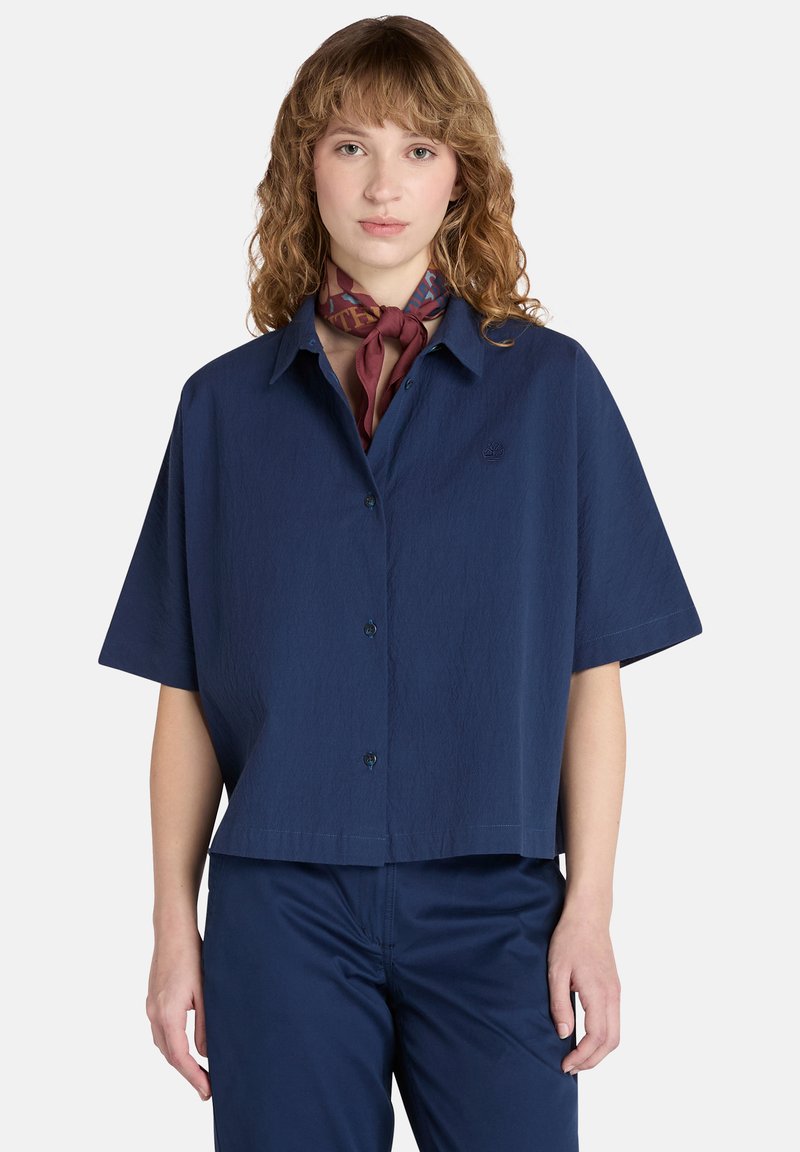 Maglietta a maniche corte blu navy con bottoni, dal taglio leggermente corto, caratterizzata da una sottile trama e un piccolo logo sul lato sinistro del torace.
