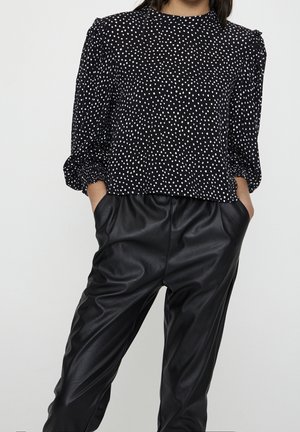 Femme portant un chemisier noir à manches longues avec des pois blancs et un pantalon en cuir noir taille haute, mains dans les poches, sur fond uni.