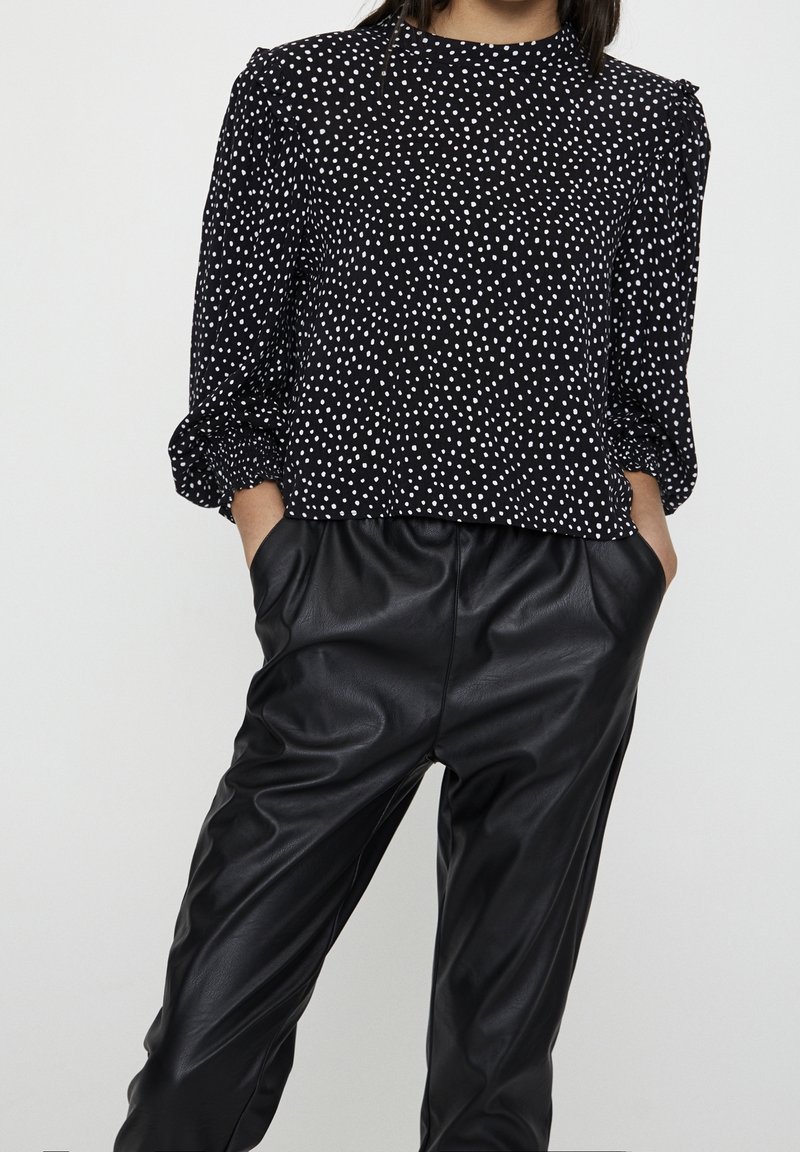 Femme portant un chemisier noir à manches longues avec des pois blancs et un pantalon en cuir noir taille haute, mains dans les poches, sur fond uni.