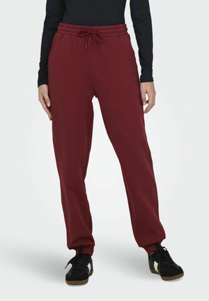 Pantalones de chándal burdeos con cinturilla elástica y cordón, con piernas ajustadas. Combinados con zapatillas negras y una camiseta de manga larga.