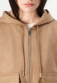 Hoodie en daim marron avec fermeture éclair sur le devant, coupe décontractée, deux poches avant et capuche réglable avec cordon. Texture douce, accents matériels minimaux.