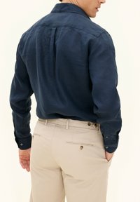 Camicia in lino blu navy con colletto classico, polsini con bottoni e piega sulla schiena; abbinata a pantaloni beige chiaro con tasche posteriori.