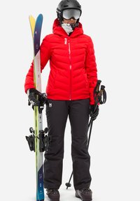 Person in roter Jacke und schwarzen Hosen hält blaue und gelbe Skier sowie Skistöcke, trägt einen schwarzen Helm und eine Schutzbrille.