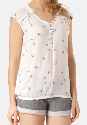 Woman wearing a white sleeveless blouse with small pink and blue floral prints and gray rolled-up denim shorts.