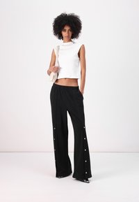 Top blanco sin mangas de corte corto y pantalones anchos negros con botones laterales, fabricados en un tejido suave y de ajuste relajado. La modelo sostiene un bolso pequeño.