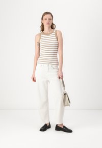 Stripet tank topp med beige og hvite striper, parret med løse hvite jeans og svarte loafers. Modellen holder en lys grå håndveske.