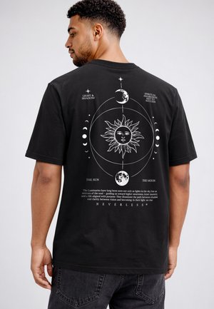 Man wearing black T-shirt with a white celestial design of sun, moon phases, and text about light, shadow, and spiritual awareness on the back.