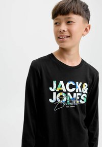 Rövid hajú fiú fülbevalóval, fekete, hosszú ujjú pólót visel, amelyen színes "JACK & JONES Essentials" felirat látható.