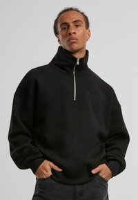 Svart zip-up hettegenser med høy krage og brodert logo, med avslappet passform, lange ermer og ribbestrikkede mansjetter.