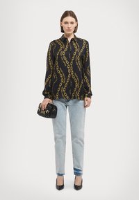 Blusa preta com estampado de correntes amarelas, mangas longas, design de botão. Usada com jeans de denim claros e uma pequena carteira preta. Saltos pretos.