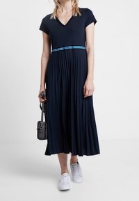 Marinblå, plisserad klänning med V-ringning och korta ärmar; har ett horisontellt blått rand vid midjan. Kombinerad med vita sneakers och en svart väska.