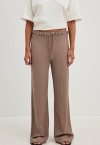 Person in locker sitzenden, gerippten taupefarbenen Hosen mit Kordelzug an der Taille und weißem, kurzärmeligen, bauchfreien Oberteil, steht vor einem schlichten hellen Hintergrund.