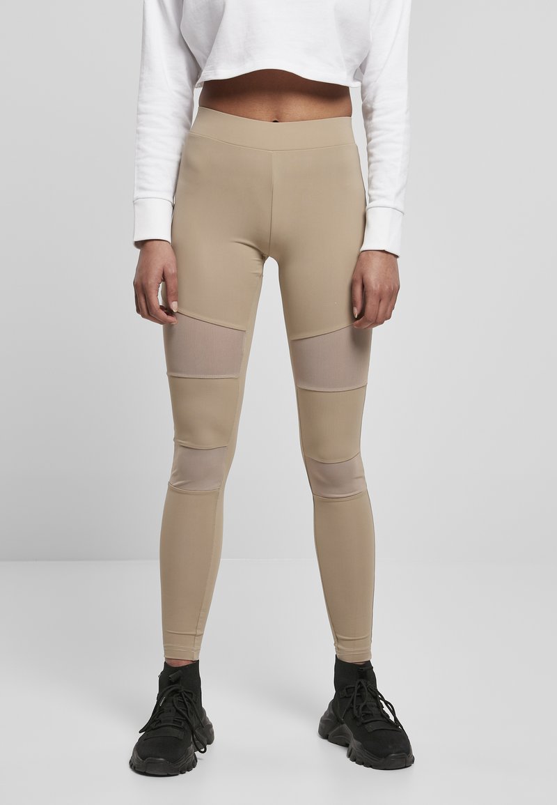 Beige Leggings mit Netzeinsätzen an den Oberschenkeln, hoher Taille und einer glatten, dehnbaren Textur. Kombiniert mit schwarzen Sportschuhen.