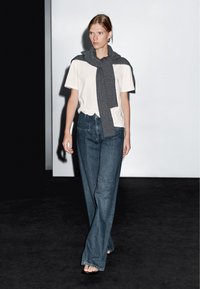 Maglietta bianca, sciarpa grigia lavorata a maglia annodata casualmente attorno alle spalle, jeans blu a vita alta e larghi, sfondo nero. Modella che cammina con sicurezza.