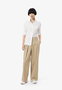 Camicia bianca con bottoni, pantaloni beige a pieghe e sneakers bianche. La camicia ha le maniche arrotolate e i pantaloni hanno una vestibilità comoda.