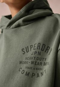 Zielona bawełniana bluza z kapturem o teksturowanym wykończeniu, z dużym czarnym napisem "SUPERDRY JPN HEAVY DUTY WORK-WEAR MFG" oraz "COMPANY."