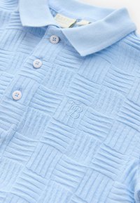 Vaaleansininen, tekstuuri-kankaasta valmistettu poolopaita, jossa on ruudullinen kuvio, kaulus ja kolme valkoista nappia. Kulmassa kirjailtu logo.