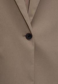 Single black button fastened on beige blazer lapel made of smooth fabric with a slight texture.
