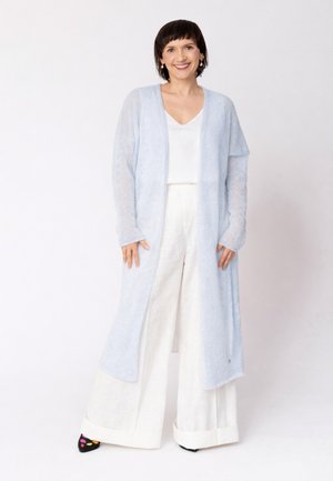 Uśmiechnięta kobieta z krótkimi ciemnymi włosami stoi na tle białego tła, ubrana w długi, jasnoniebieski kardigan, biały top, szerokie białe spodnie i wzorzyste buty.